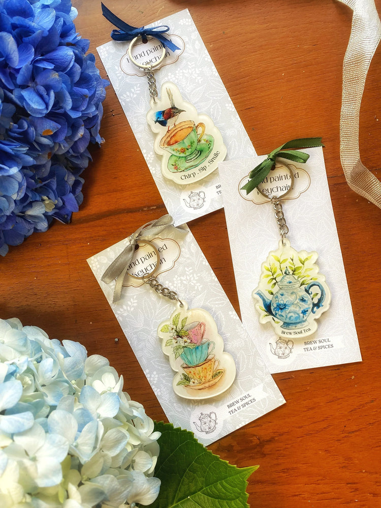 Hand-painted tea-themed keychains with teacup and teapot designs on wooden table, surrounded by blue hydrangea flowers.