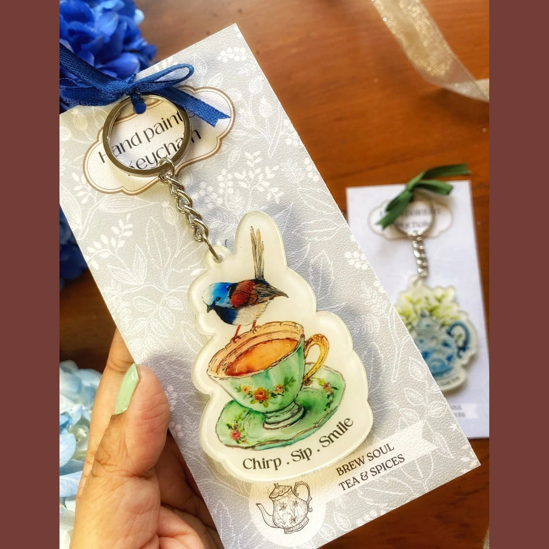 Hand-painted keychain featuring a colorful bird perched on a floral teacup with 'Chirp. Sip. Smile.' text, displayed on decorative cardstock.