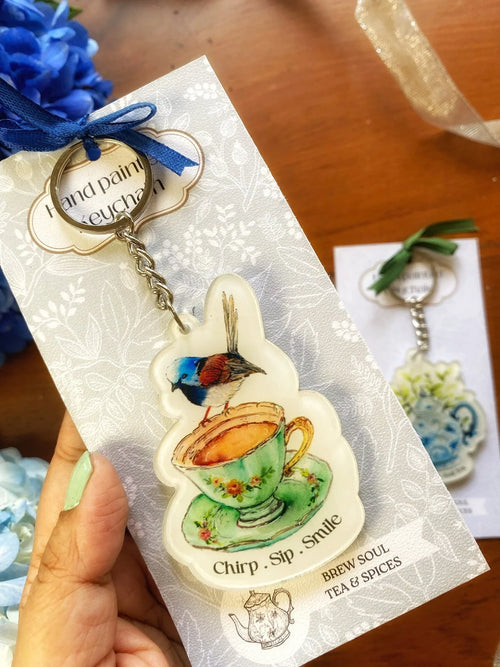 Hand-painted keychain featuring a colorful bird perched on a floral teacup with 'Chirp. Sip. Smile.' text, displayed on decorative cardstock.