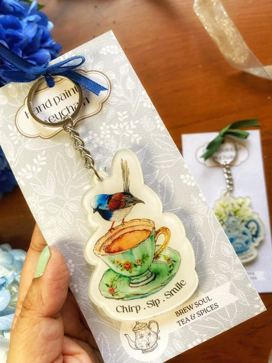 Hand-painted keychain featuring a colorful bird perched on a floral teacup with 'Chirp. Sip. Smile.' text, displayed on decorative cardstock.